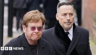 Elton John, who has short gingery hair and wears purple-tinted spectacles, pictured walking on a London street with his husband David Furnish, who has short grey hair. Both have serious expressions
