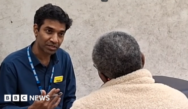 A man is sitting down and is wearing a navy blue shirt, open at the neck, and wearing a blue lanyard with NHS in white letters. He's holding a pen in his hand and is speaking to an older woman, with grey hair and glasses. We can only see the back of her head as she's facing him, she's wearing a beige fleece coat with the collar pulled up to protect her neck.