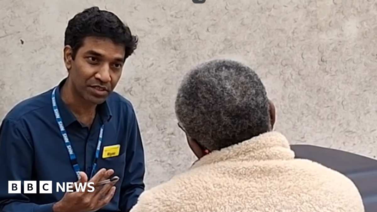 A man is sitting down and is wearing a navy blue shirt, open at the neck, and wearing a blue lanyard with NHS in white letters. He's holding a pen in his hand and is speaking to an older woman, with grey hair and glasses. We can only see the back of her head as she's facing him, she's wearing a beige fleece coat with the collar pulled up to protect her neck.