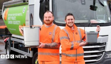 Two men in orange jump suits with Chichester District Council logos on them. They are smiling and holding small bins. They are standing in front of a lorry.