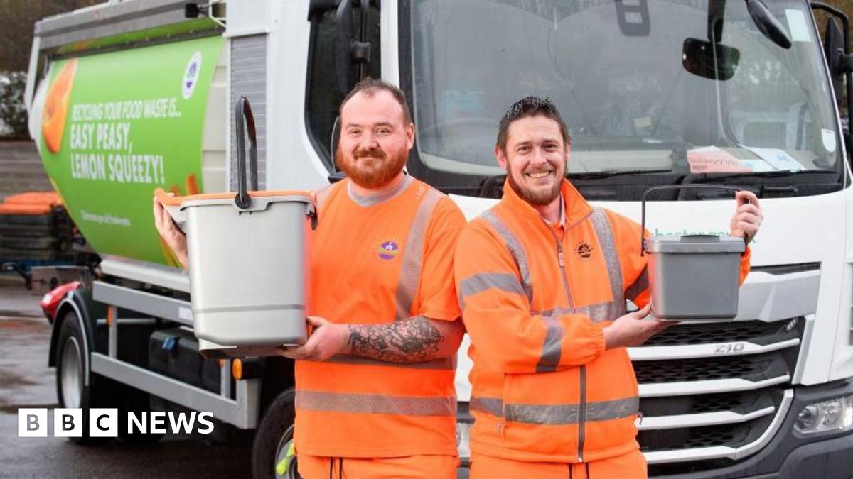 Two men in orange jump suits with Chichester District Council logos on them. They are smiling and holding small bins. They are standing in front of a lorry.