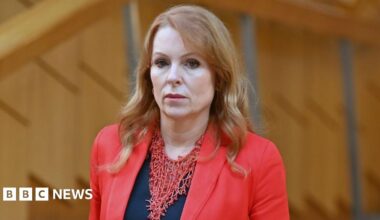 Ash Regan, who has long ginger hair, looks towards the camera, with wood panelling behind her. She is wearing a red jacket over a black top.