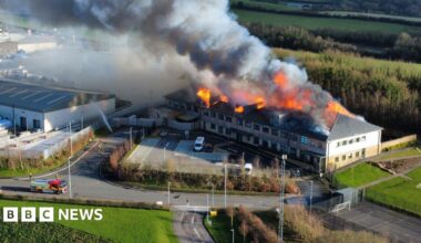 A drone image of a school building which is on fire, with a big cloud of smoke coming out of the roof of the building.There is a fire truck and firefighters in the left of the photo.