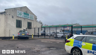 A large light brown building with a sloped roof and metal railings at the front and a police car parked in the foreground