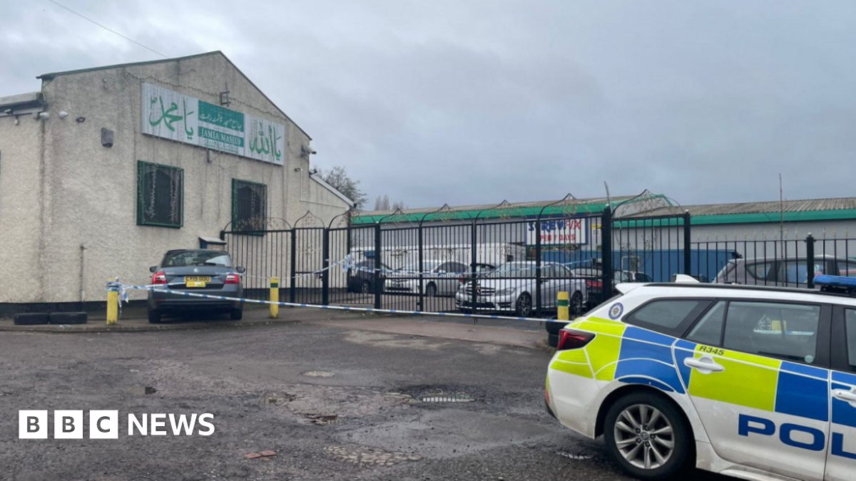 A large light brown building with a sloped roof and metal railings at the front and a police car parked in the foreground