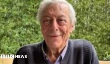 An elderly man with grey hair and wearing a dark blue jumper smiles at the camera