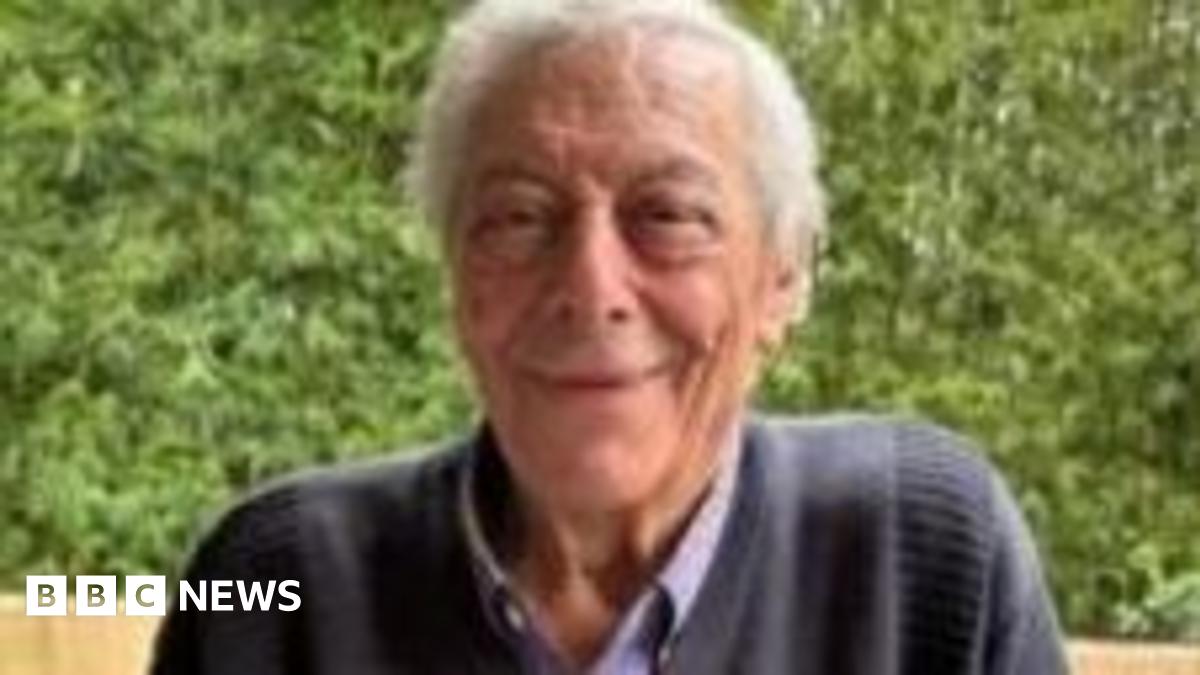 An elderly man with grey hair and wearing a dark blue jumper smiles at the camera