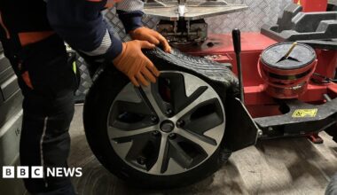 Part of the road with large, wavy cracks in the tarmac and raised sections. Orange and white cones stretch along the middle of the road as cars pass.