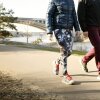 Two people wearing sneakers and photographed from the neck down walk on a paved path that runs along a body of water. A bridge is in the background.