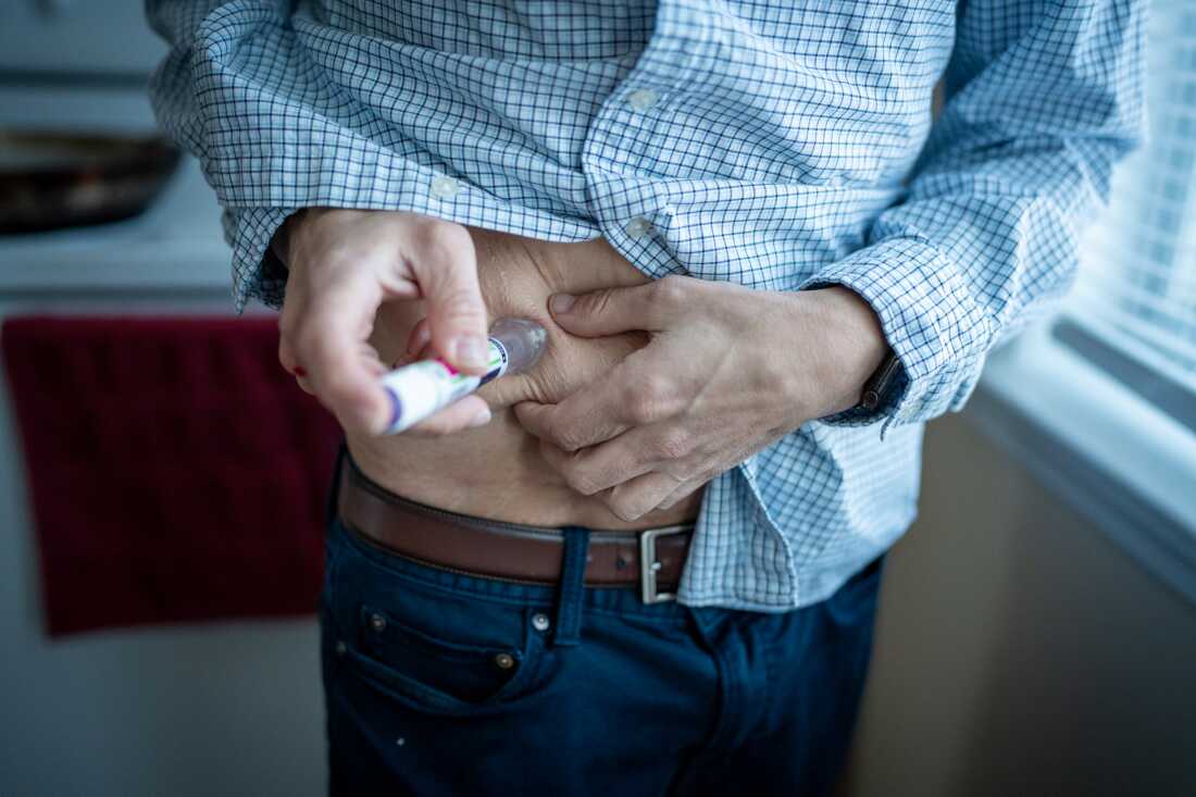 Jason Krynicki lifts a blue-checked shirt and pinches the skin of his abdomen to give himself his GLP1 injection.