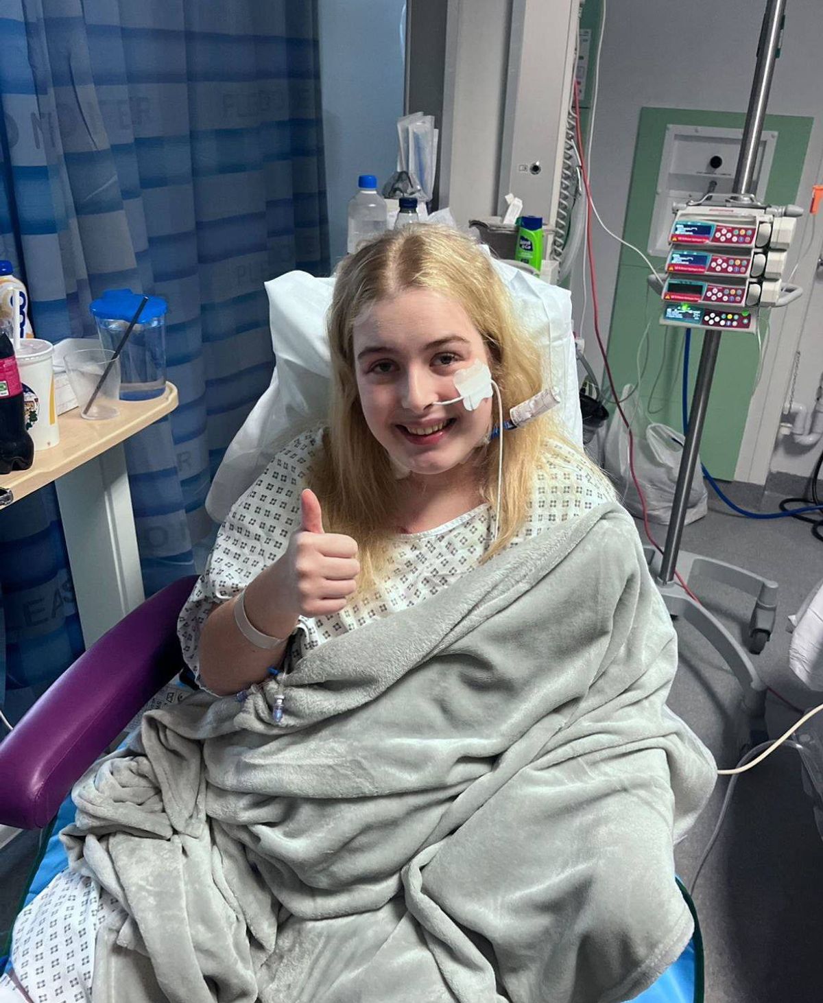 An individual in a hospital gown, appearing to be in a positive mood, is giving a thumbs-up gesture while seated in a hospital bed. Various medical devices and a table with objects are visible in the background.