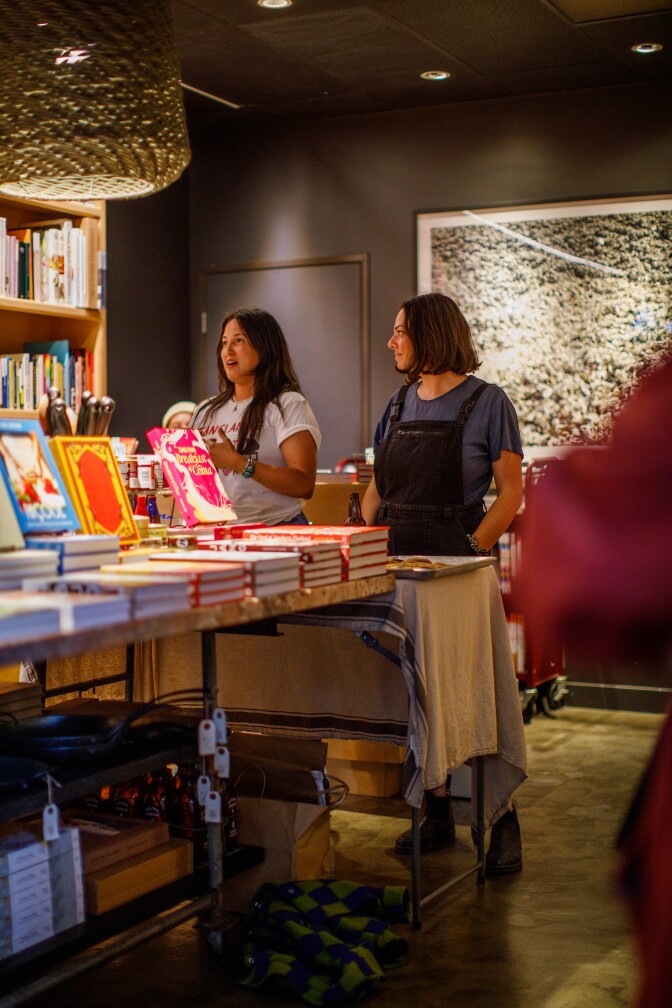 Two women, light and medium skinned, stand in bookstore surrounded by books. One has long brown hair; the other shoulder length hair. One is speaking, the other is standing looking at her