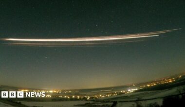 A photograph using 30-second exposure  showing the Falcon 9 upper stage re-entering the atmosphere above Berlin, Germany. A long streak of bright yellow and white is seen in the dark night sky, above a landscape of dark field and bright lights from the city.