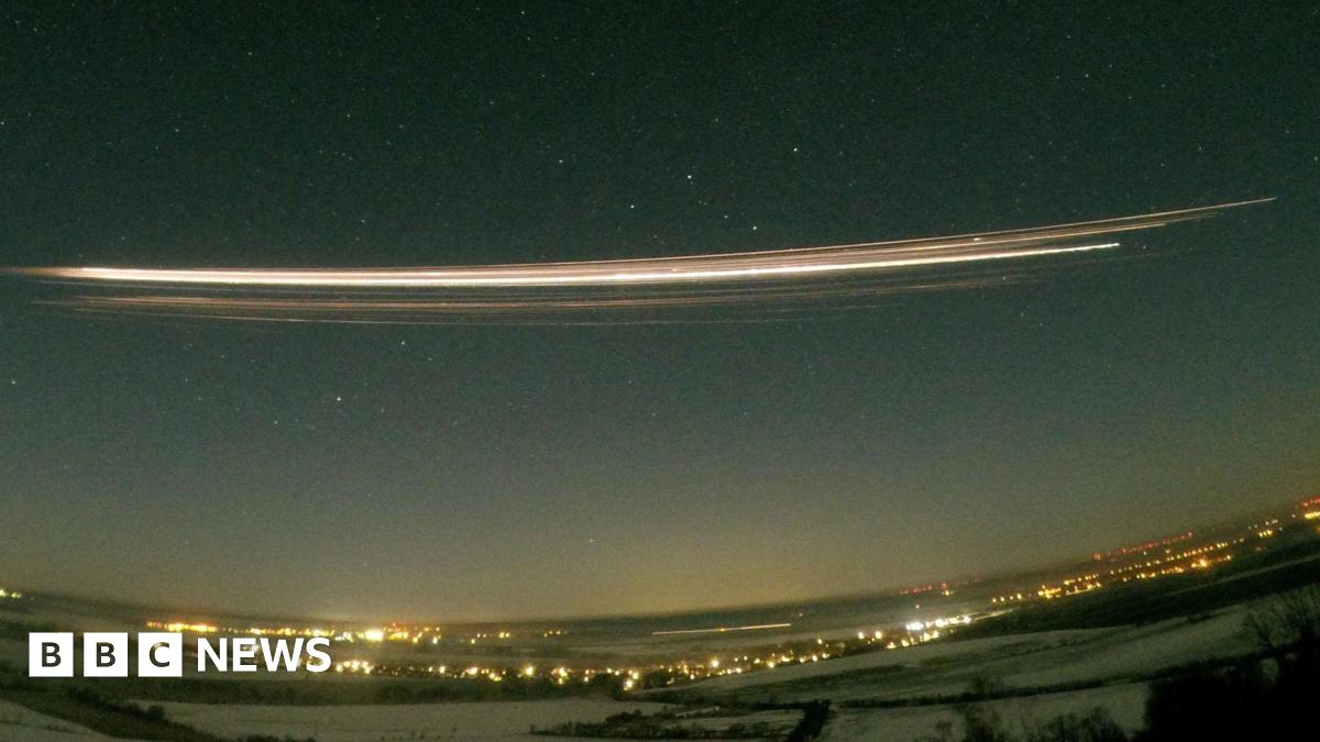 A photograph using 30-second exposure  showing the Falcon 9 upper stage re-entering the atmosphere above Berlin, Germany. A long streak of bright yellow and white is seen in the dark night sky, above a landscape of dark field and bright lights from the city.