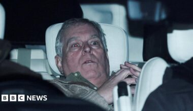 Andrew Mountbatten-Windsor is slumped in the back seat of a car with a white headrest. He looks shocked and has grey hair. He is wearing a green jumper and his hands are steepled together. His eyes are wide and the flash has turned them red.