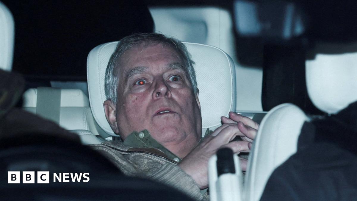 Andrew Mountbatten-Windsor is slumped in the back seat of a car with a white headrest. He looks shocked and has grey hair. He is wearing a green jumper and his hands are steepled together. His eyes are wide and the flash has turned them red.