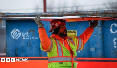 A bearded construction worker wearing an orange sweatshirt and yellow safety vest carries a sheet of construction material over his his head.  In the background there is a a blue container. The picture was taken at the site of the Gateway Tunnel construction project as seen from North Bergen, New Jersey, on February 10, 2026.