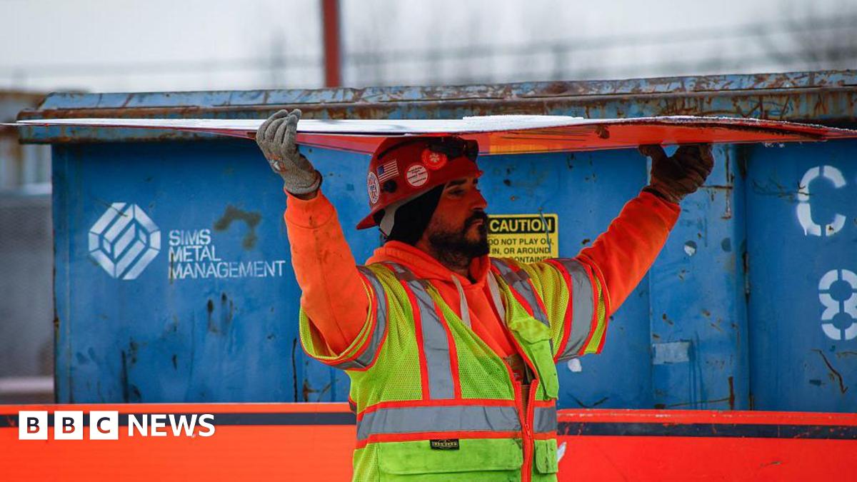 A bearded construction worker wearing an orange sweatshirt and yellow safety vest carries a sheet of construction material over his his head.  In the background there is a a blue container. The picture was taken at the site of the Gateway Tunnel construction project as seen from North Bergen, New Jersey, on February 10, 2026.