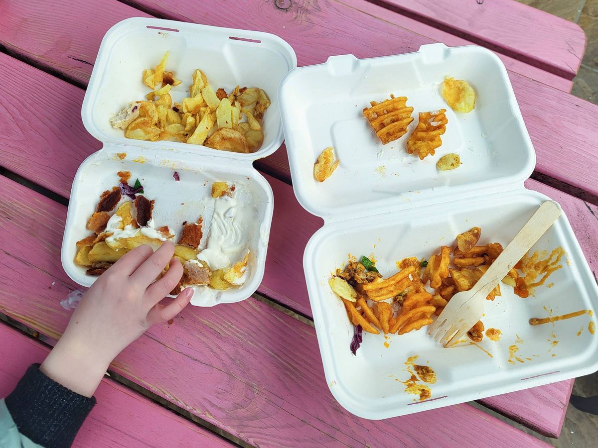 Three rectangular white Styrofoam containers, partially filled with remnants of a meal, are positioned on a pink wooden table. A hand, partially visible, holds one of the containers, suggesting recent consumption.