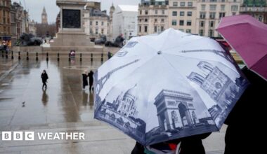 Two people in the foreground stand under open umbrellas as rain falls in front of a monument in London