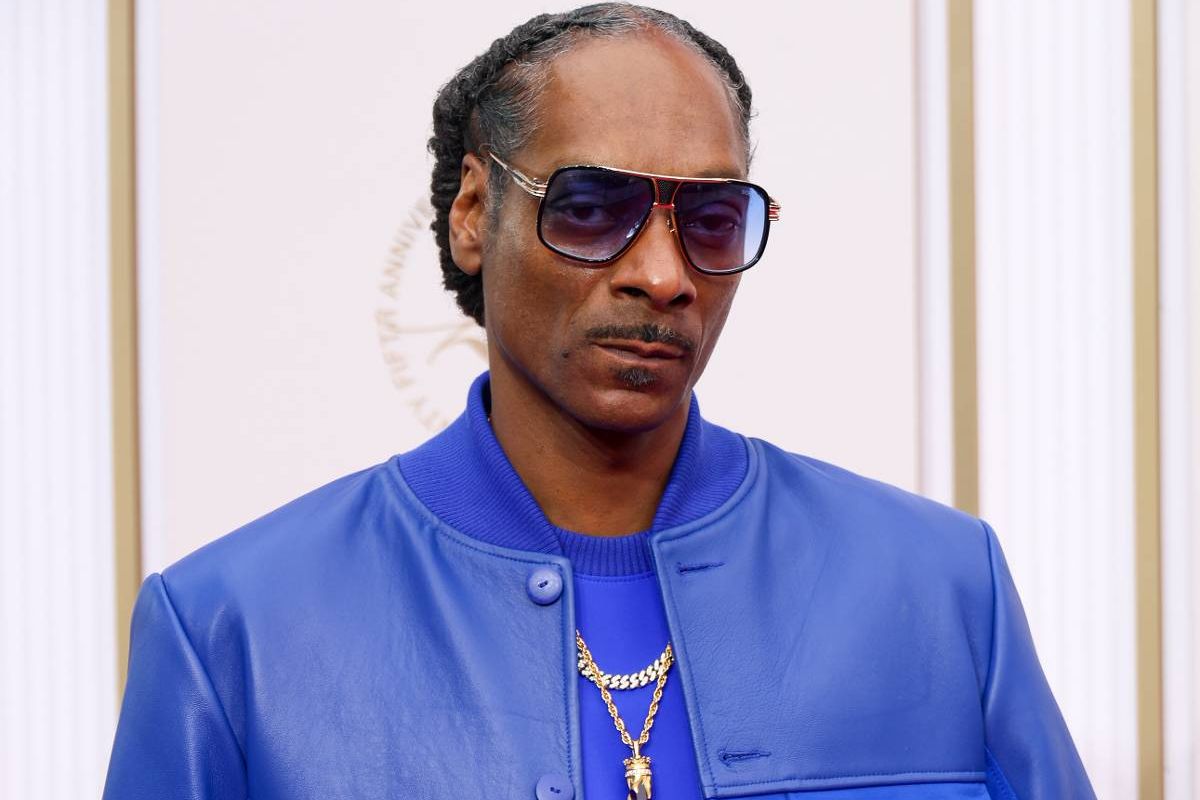 An individual dressed in a blue outfit stands facing the camera with a serious expression, wearing sunglasses.