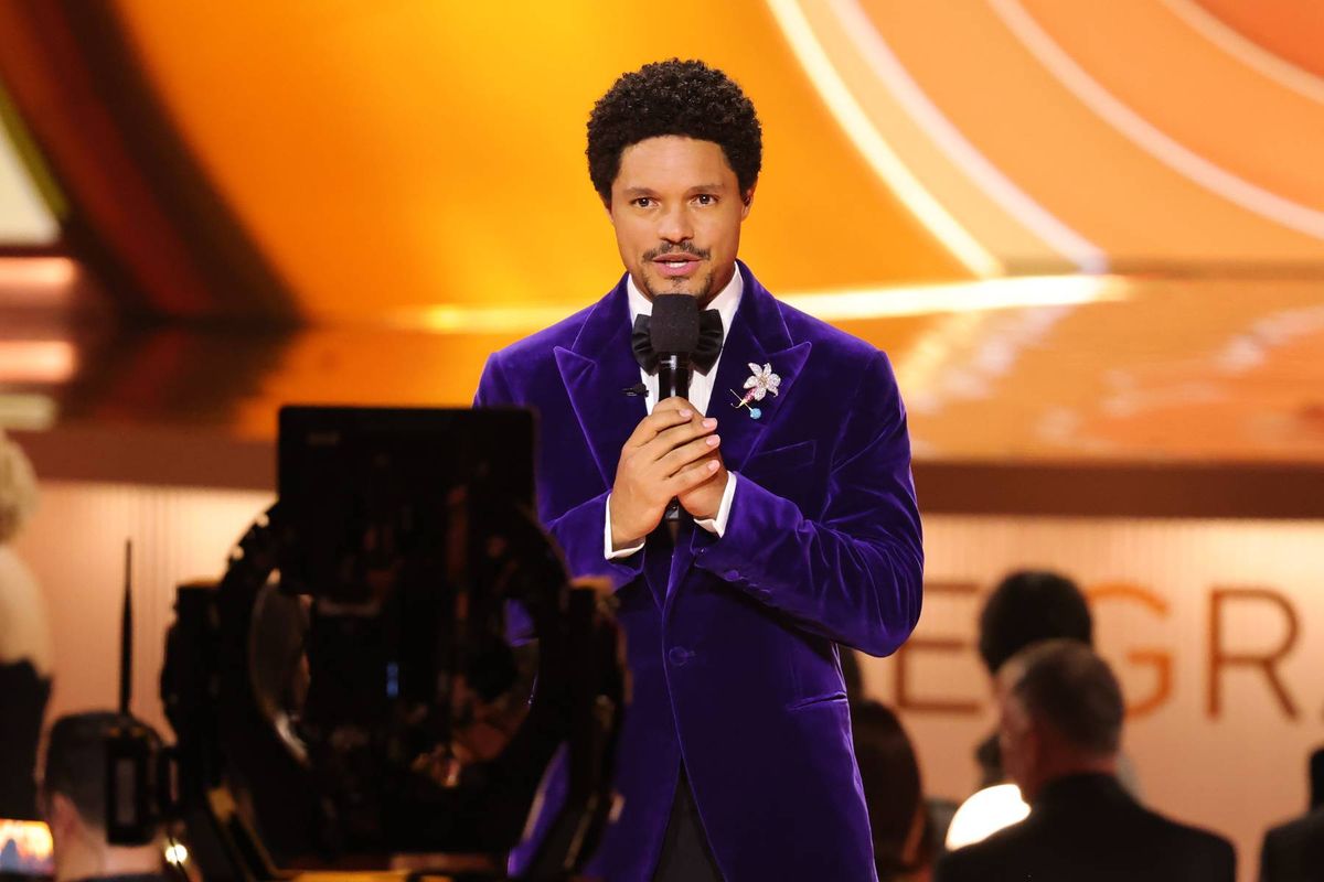 An individual dressed in a vibrant purple suit stands on a stage, holding a microphone, and addressing an audience, with a blurred background featuring seated spectators and a partially visible banner.