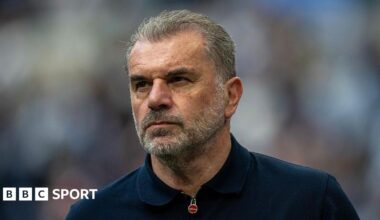 Close-up of Ange Postecoglou. He has short grey hair, brushed away from his face, and a grey beard. He is wearing a navy top which has a zip fastening at the collar.