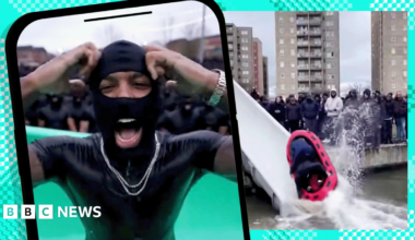 A composite image showing a graphic of a phone with an image of a young black man wearing a black balaclava and a gold chain shouting and raising his hands. In the background is a still from an AI video showing a waterslide with a red dinghy sliding down it into dirty water, with tower blocks and a crowd of young men in hoodies and balaclavas.