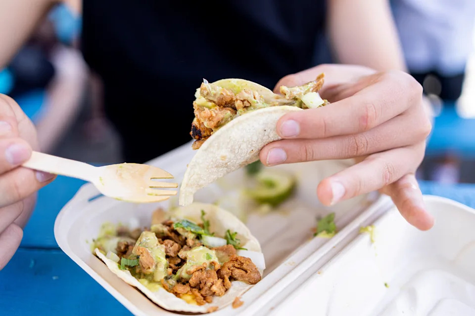 Person eating tacos with a fork, holding one taco filled with toppings. Plate holds additional taco. Casual dining setting