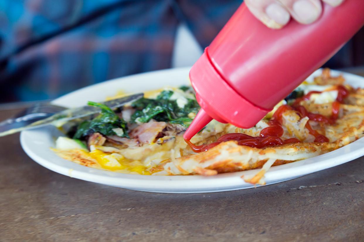 A man sits at a restaurant table, squeezing ketchup out of a bottle onto his breakfast.