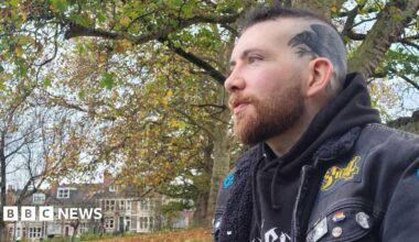 Patrick Darling wearing a black hoodie underneath a black denim jacket, decorated with various colourful pins and patches. He has dark hair which is shaved on the sides, and a tattoo of a raven across the left side of his head. He is sitting outdoors in a park surrounded by trees, looking off to the left of the camera. In the background there is a row of terraced houses.