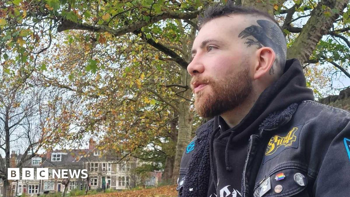 Patrick Darling wearing a black hoodie underneath a black denim jacket, decorated with various colourful pins and patches. He has dark hair which is shaved on the sides, and a tattoo of a raven across the left side of his head. He is sitting outdoors in a park surrounded by trees, looking off to the left of the camera. In the background there is a row of terraced houses.