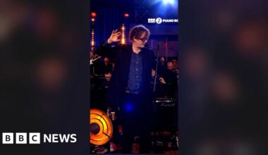 A man, Jarvis Cocker, in a blue shirt and suit, with one hand raised and outstretched, looking to the side.