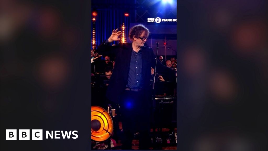 A man, Jarvis Cocker, in a blue shirt and suit, with one hand raised and outstretched, looking to the side.