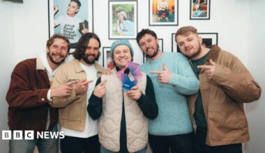 The band hold up their trophy for topping the download chart, which is shaped like a number one. Famous singers are in framed pictures behind them, including Robbie Williams.