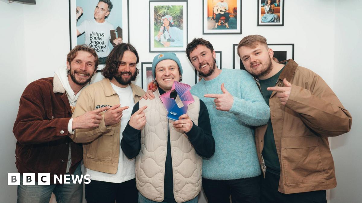 The band hold up their trophy for topping the download chart, which is shaped like a number one. Famous singers are in framed pictures behind them, including Robbie Williams.