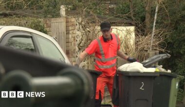A man wearing pink high visibility clothing. He is wearing black gloves and a black cap. There are three black bins around him.
