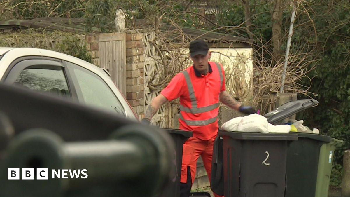 A man wearing pink high visibility clothing. He is wearing black gloves and a black cap. There are three black bins around him.