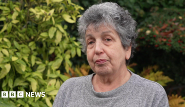 A woman with grey hair wearing a grey jumper is standing outside. Bushes are behind her filling most of the background.