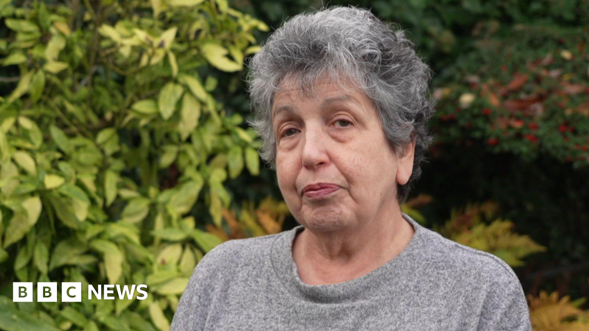 A woman with grey hair wearing a grey jumper is standing outside. Bushes are behind her filling most of the background.