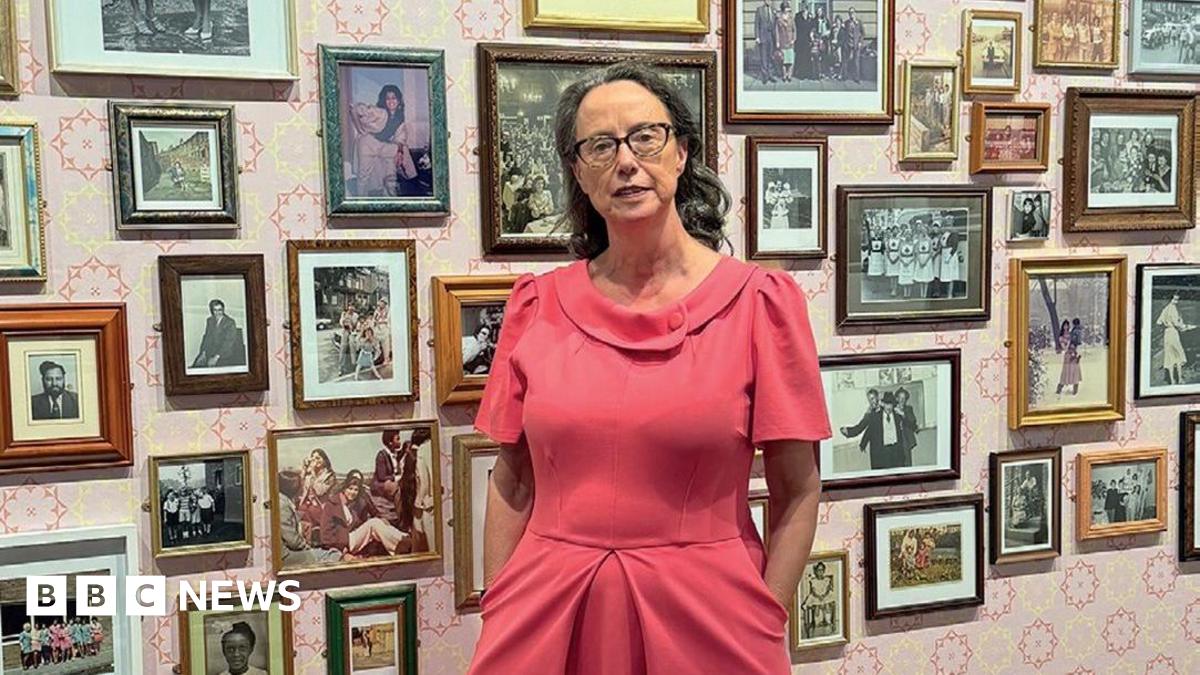 A woman in a pink dress stands against a backdrop of small photographs on a pink and white wall