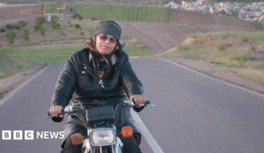 A woman rides a motorcycle along an empty road, with rolling hills seen in the background.