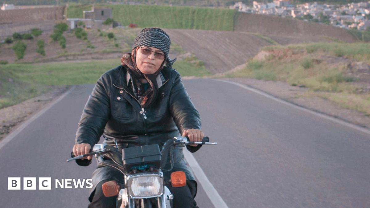A woman rides a motorcycle along an empty road, with rolling hills seen in the background.