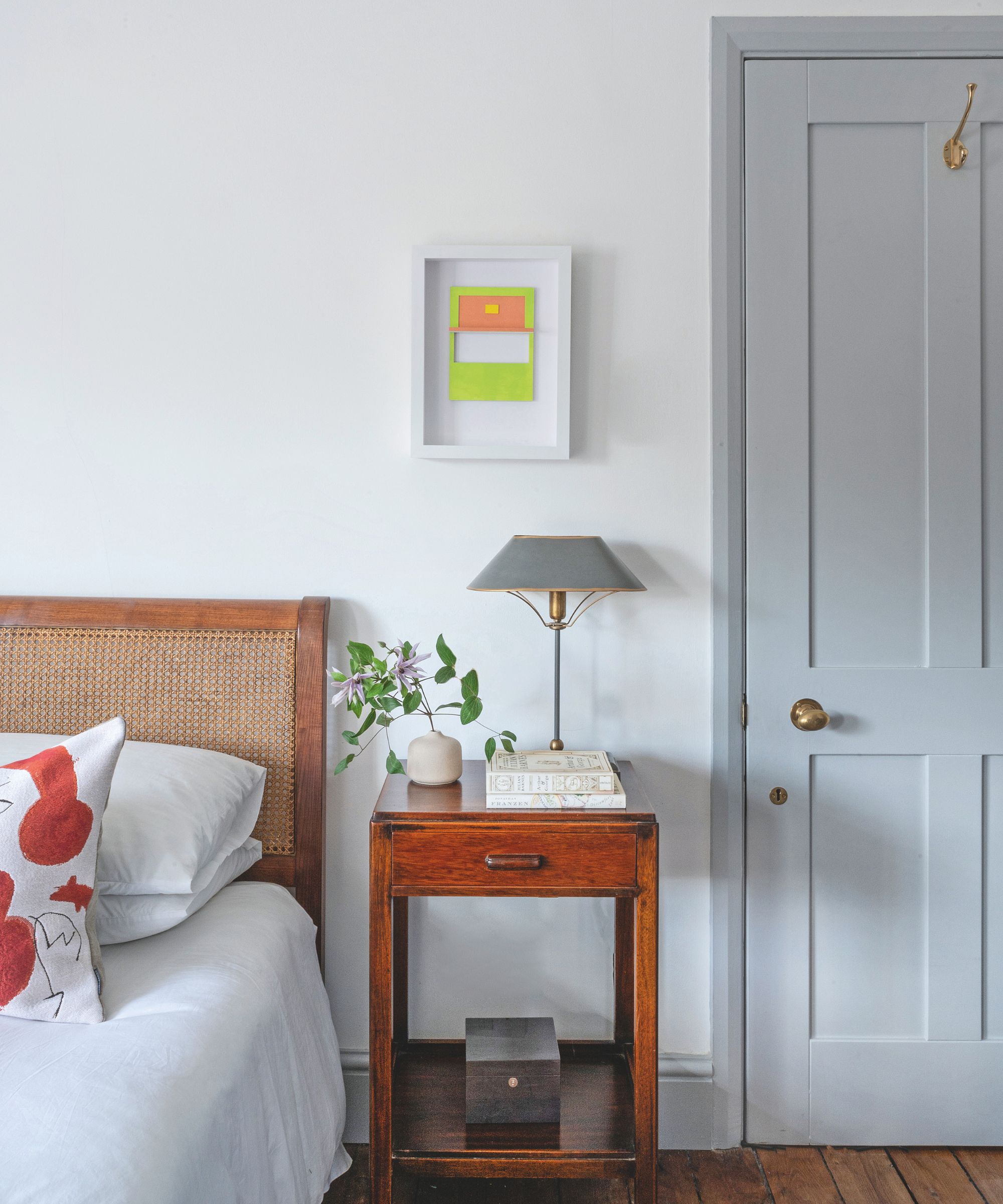A minimalist bedroom featuring a bed with a woven cane headboard, a dark wood nightstand, and a light gray door