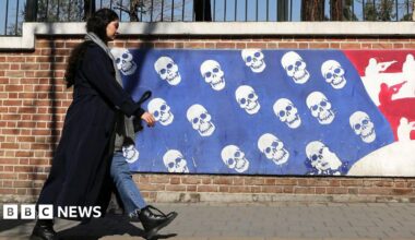 A woman in black in Iran walks near a mural that depicts the US flag with skulls instead of stars