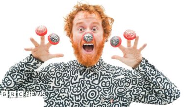 A man with red hair and beard is dressed in a black and white top, featuring cartoon drawings of a variety of characters. He is wearing one red nose with black and white doodle illustrations, and four of his fingers with a red nose with black and white doodle illustrations.
