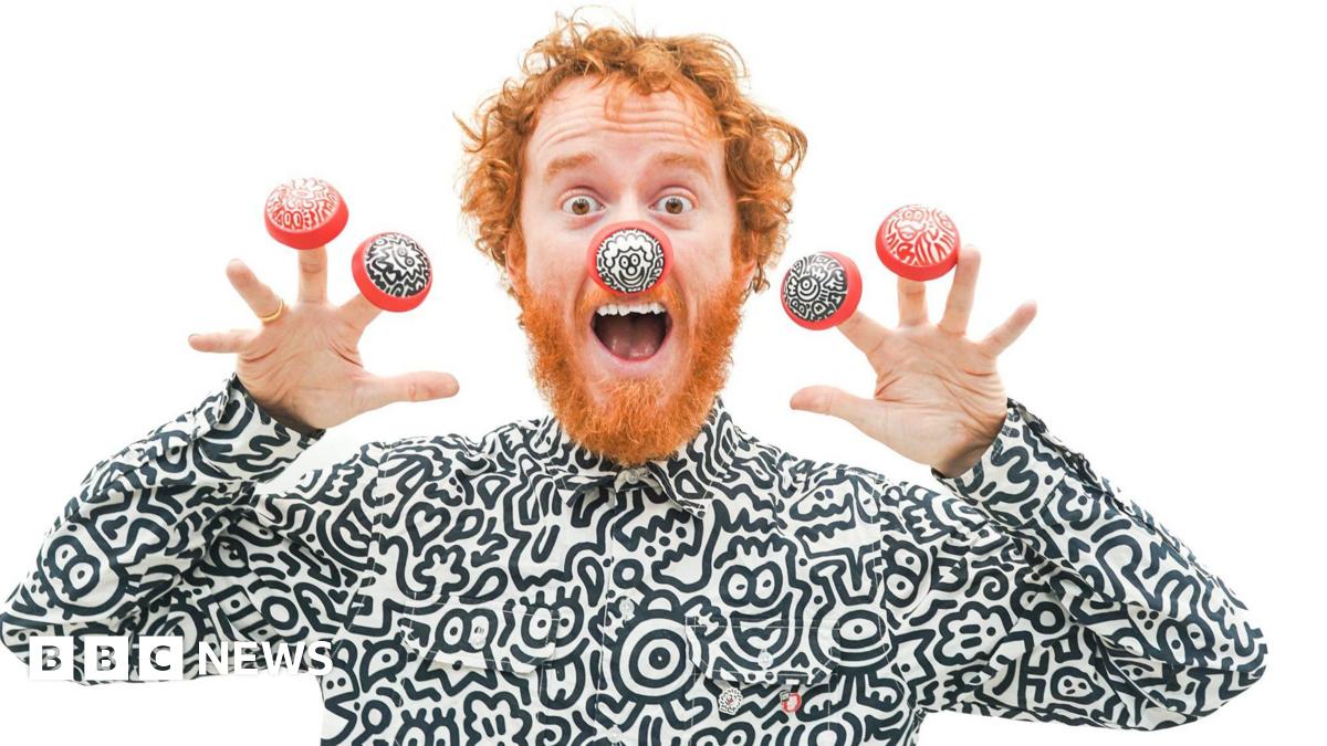 A man with red hair and beard is dressed in a black and white top, featuring cartoon drawings of a variety of characters. He is wearing one red nose with black and white doodle illustrations, and four of his fingers with a red nose with black and white doodle illustrations.