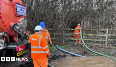 Three men in orange high visibility suits position hoses into muddy water in a woodland.