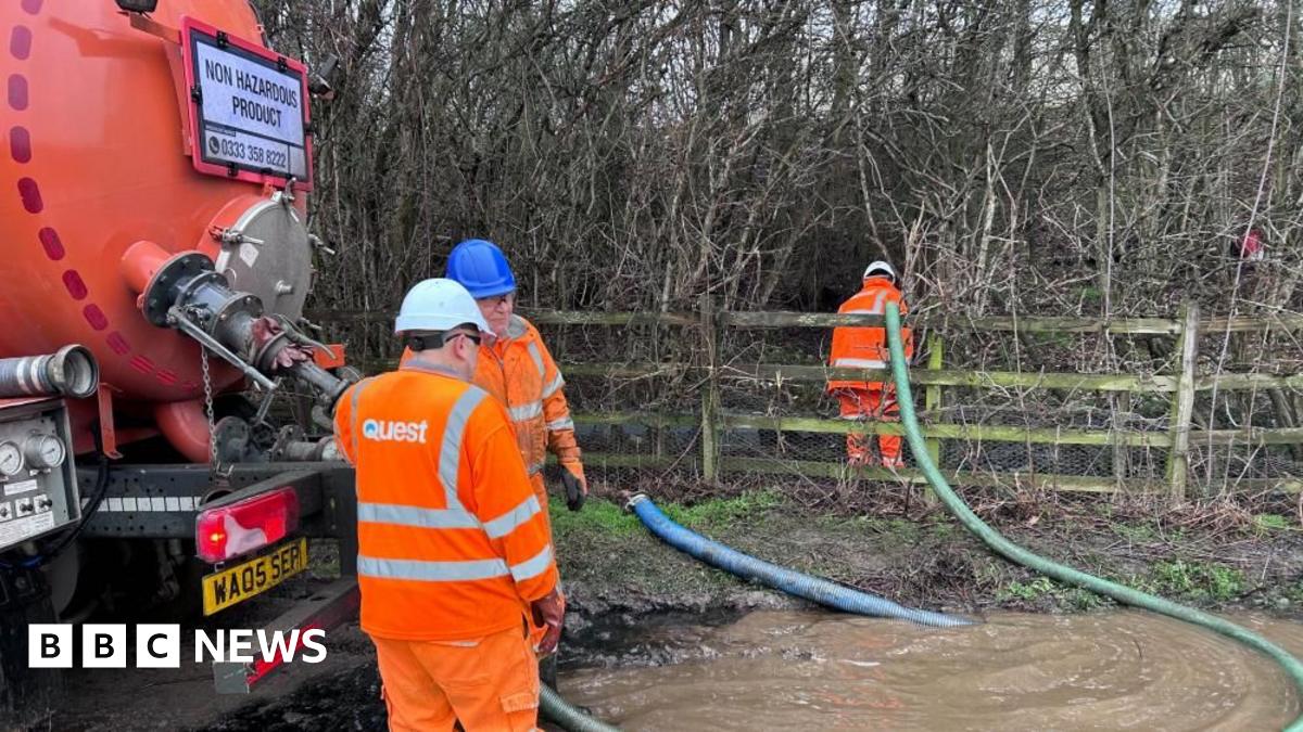 Three men in orange high visibility suits position hoses into muddy water in a woodland.