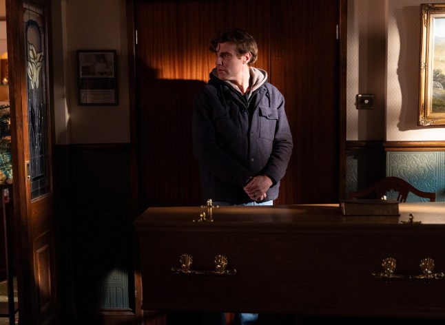 Theo stands alone in the funeral parlour beside a coffin in Corrie